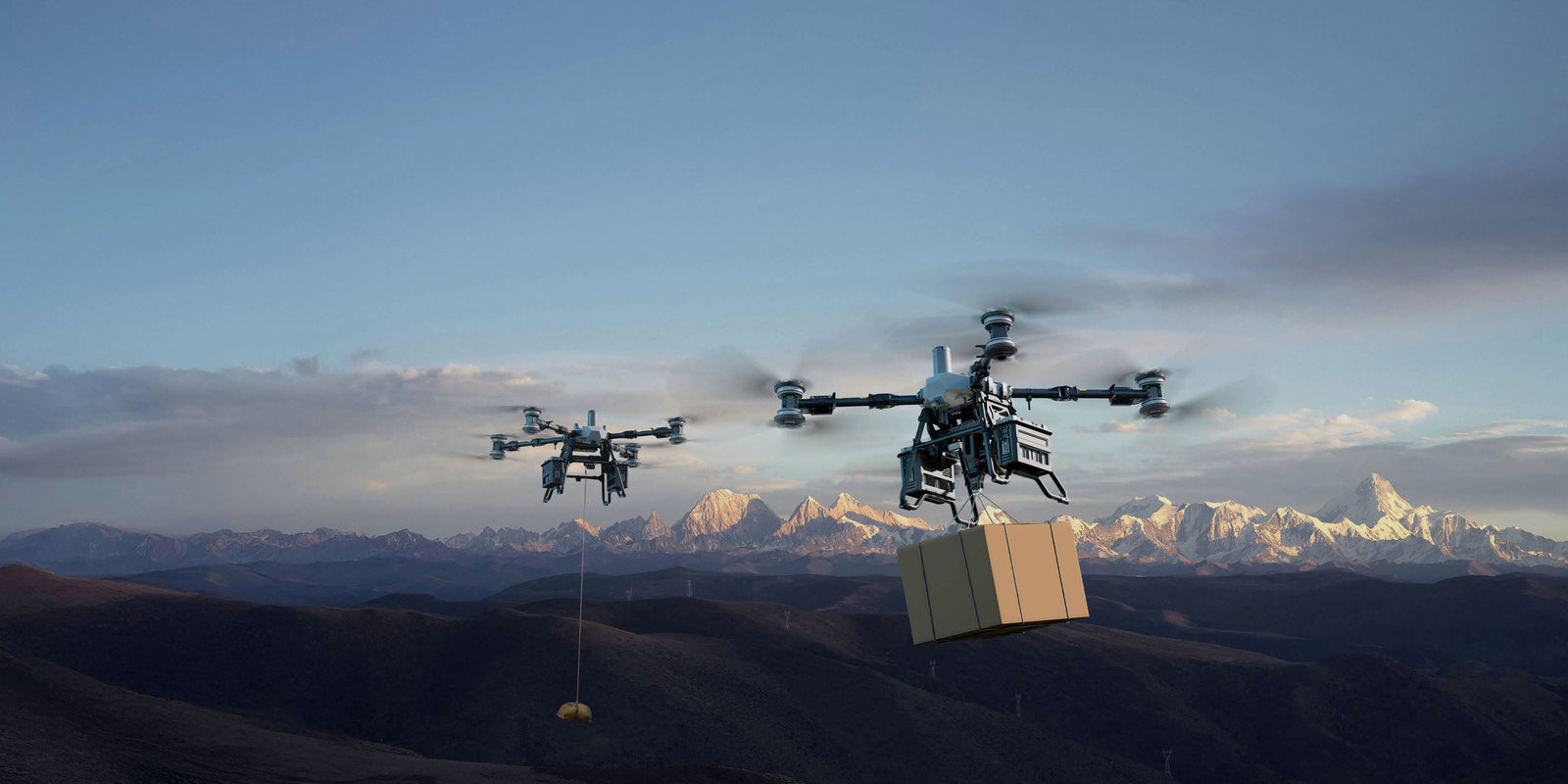 Two DJI FlyCart 100 drones flying with packages against a mountainous backdrop at sunset.